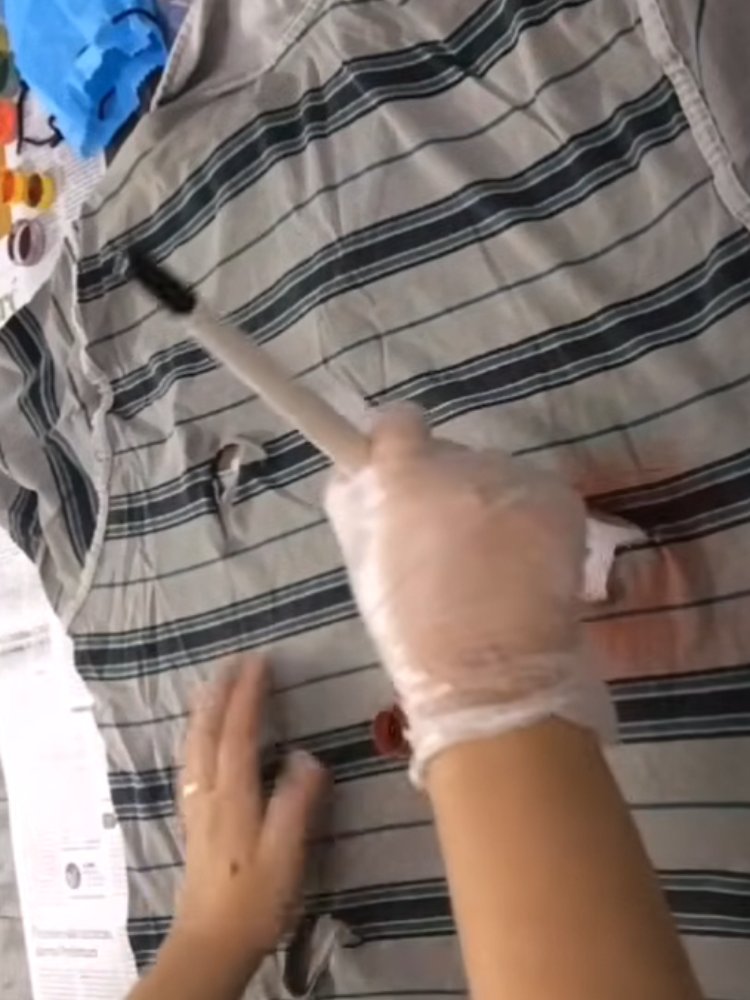 Mulher de pele clara com luvas pintando camiseta rasgada cinza e verde com tinta vermelha 