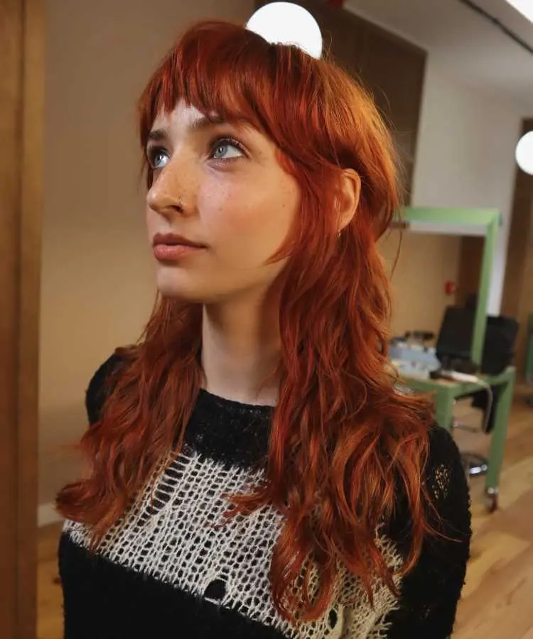 Mulher com cabelos longos e ruivos, estilizados em corte mullet com franja curta e desfiada. Ela veste uma blusa preta com detalhes rendados, olha para cima com expressão contemplativa, em um ambiente interno com iluminação quente e moderna.