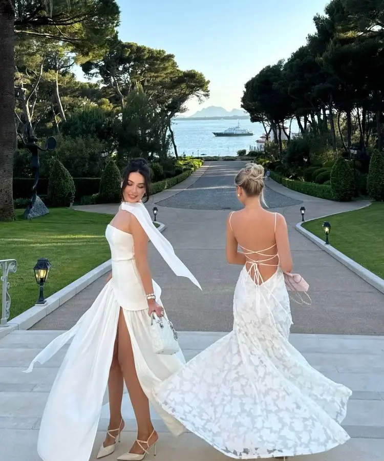Duas mulheres em vestidos longos brancos descem uma escadaria de pedra em um jardim elegante. Elas estão viradas, com uma delas olhando por cima do ombro. O cenário inclui árvores, gramados e uma vista para o mar com um iate.