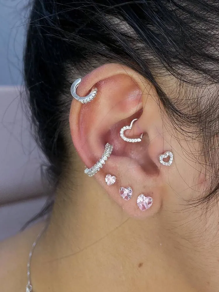 Orelha com piercing na hélix, argola de coração no daith, coração no tragus, argola no conch e três brincos pequenos no lóbulo.