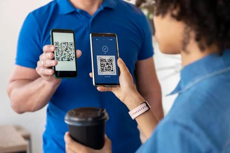 Homem de blusa azul segurando celular que exibe QR Code enquanto uma mulher também vestida de azul segura um celular lendo esse QR Code. Além do celular, ela também segura um copo preto de café.