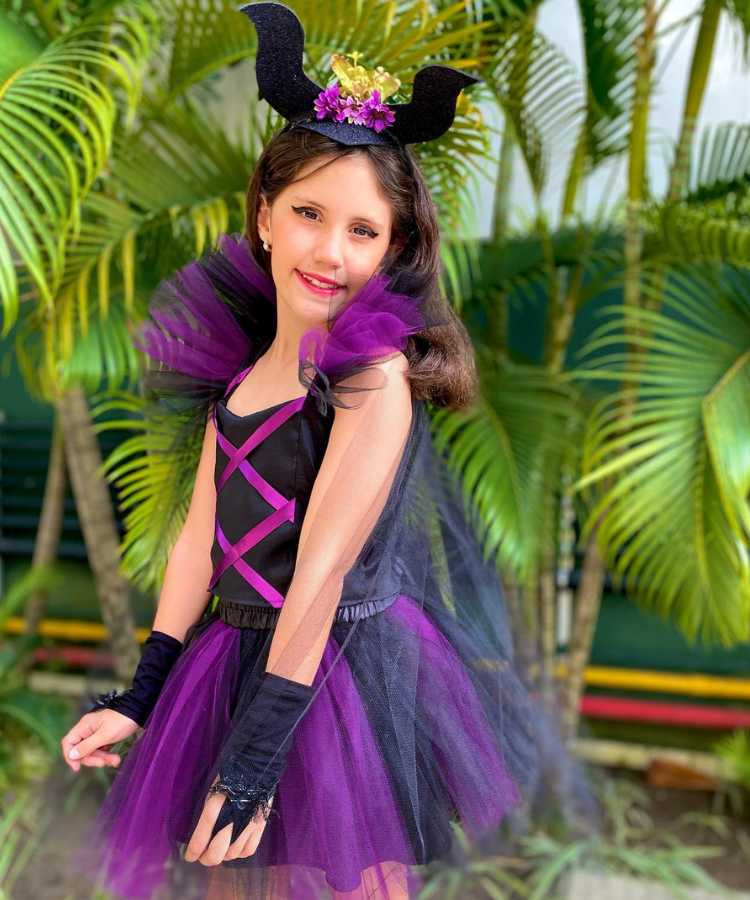 Garota com fantasia de bruxa infantil Malévola. Cores predominantes: preto e roxo. Usa: vestido, tiara de chifres em E.V.A., luva