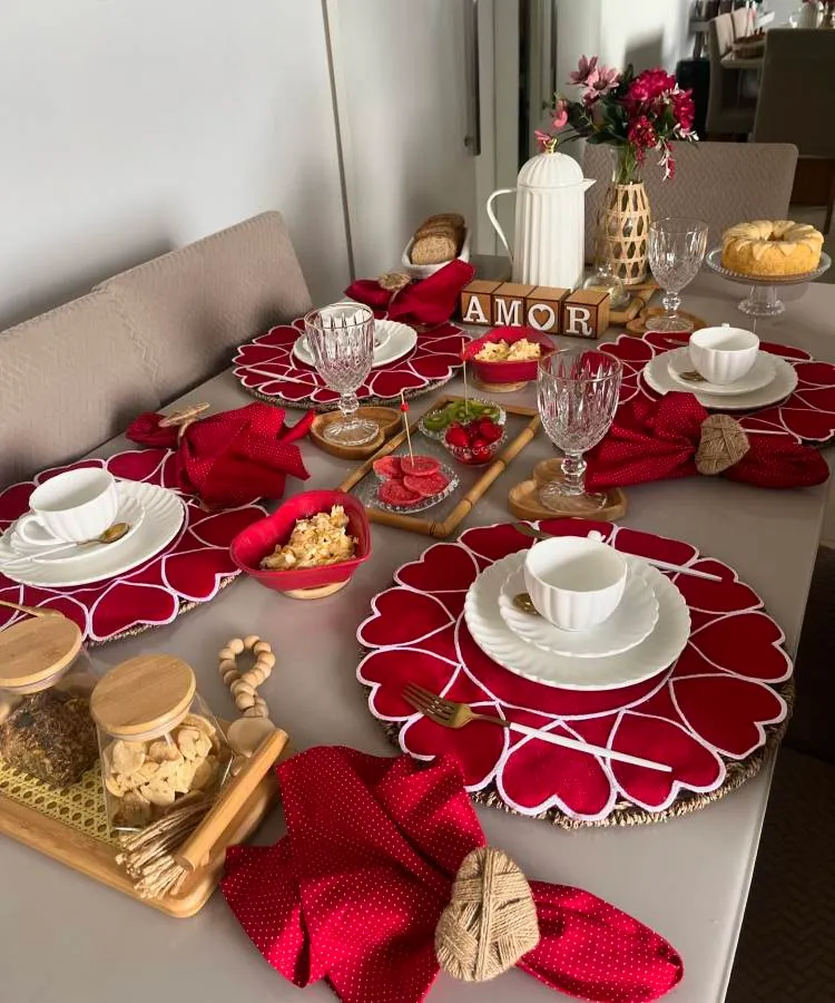 A imagem exibe uma mesa posta para café da manhã, destacando sousplats vermelhos em formato de coração. Pratos brancos e xícaras estão acompanhados de guardanapos vermelhos com porta-guardanapos de palha. Taças de vidro elegante completam a arrumação. No centro, há uma garrafa térmica branca e blocos de madeira formando a palavra "AMOR". Bandejas de madeira contêm biscoitos e pães, enquanto um prato elevado exibe um bolo. Um vaso com flores vermelhas adiciona charme ao ambiente, criando um cenário romântico e acolhedor.


