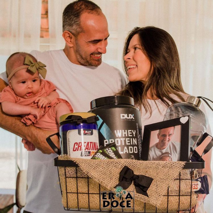Foto de homem segurando bebê e mulher segurando cesta com whey protein, creatina e porta-retrato