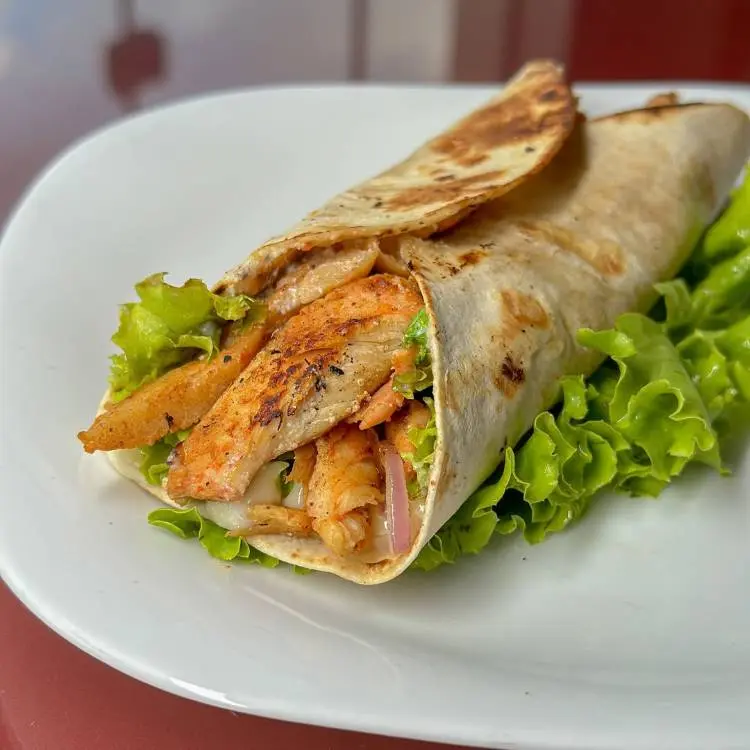 Wrap de frango grelhado suculento, recheado com alface fresca e cebola roxa, todo enrolado em um pão macio e dourado. O prato é apresentado em um prato branco sobre uma superfície vermelha, destacando os ingredientes saborosos e frescos.