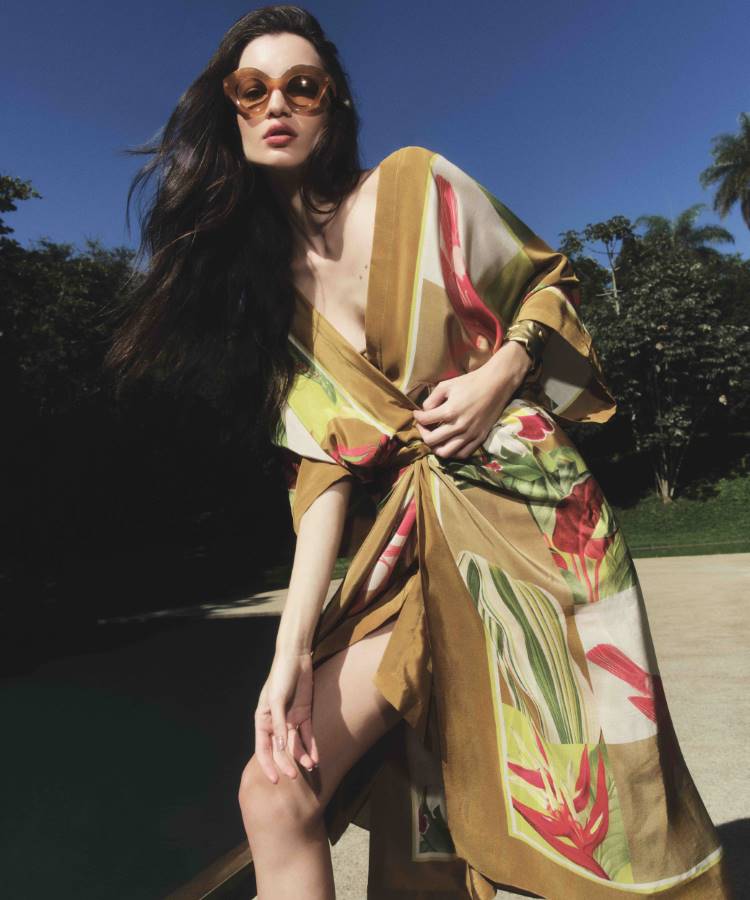 Mulher de cabelos escuros e óculos de sol posa ao ar livre, vestindo um vestido solto de seda com estampa floral em tons de marrom, amarelo e vermelho. O vestido tem uma amarração na frente.