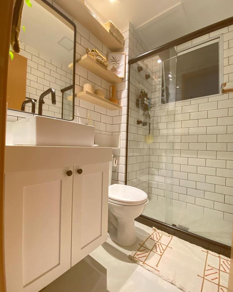 Banheiro branco com luz amarela, plantas, pia branca, espelho, vaso sanitário, decorações em tons terrosos e prateleiras