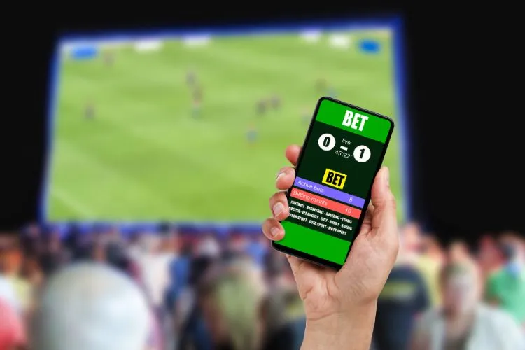 Uma mão segura um smartphone exibindo um aplicativo de apostas com resultados de jogo, possivelmente simulando o Bet365. Ao fundo, uma partida esportiva é projetada para uma multidão, ilustrando o engajamento com apostas online e a potencial ligação com a ludopatia.