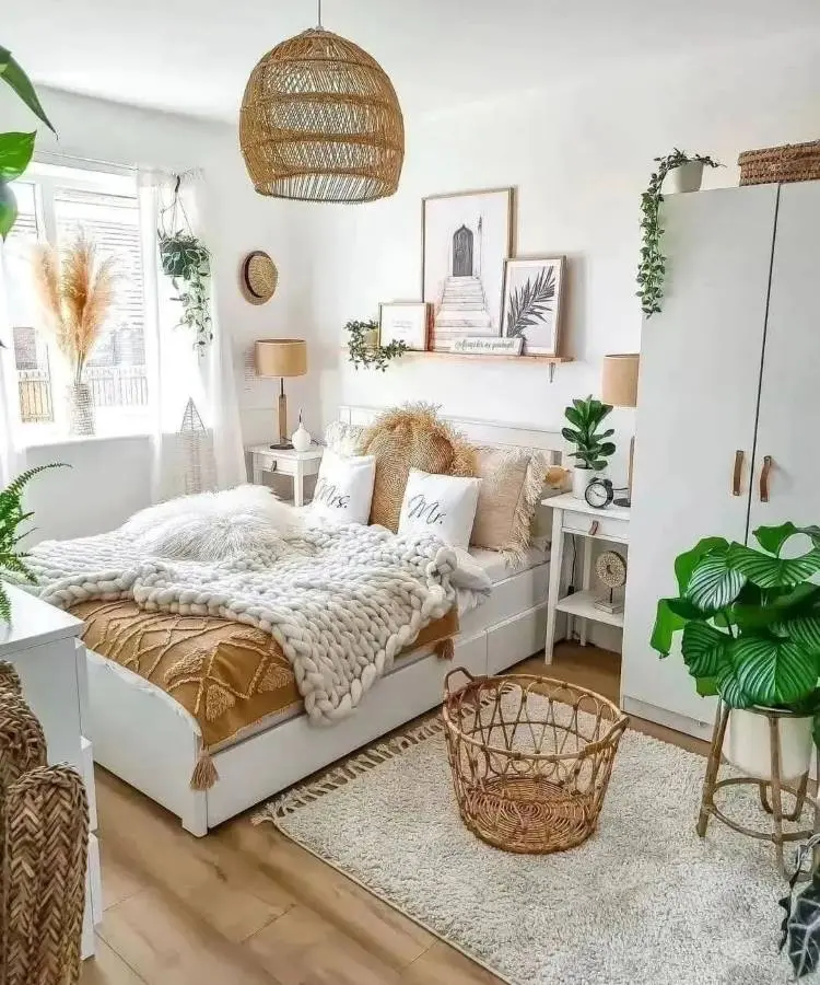 Quarto de casal branco, marrom palha e verde, cama com almofadas, cobertores, paredes decoradas com quadros, plantas e objetos de palha