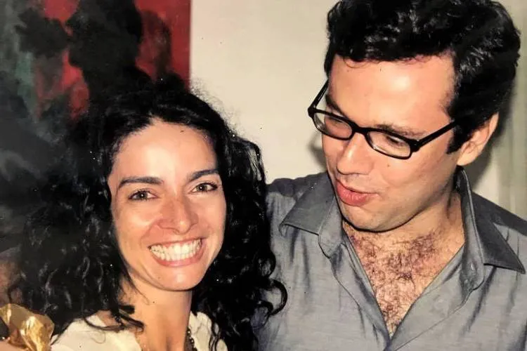 Uma foto antiga mostra uma mulher com cabelo escuro e encaracolado sorrindo amplamente ao lado de um homem de óculos. Ele usa uma camisa cinza e tem o braço em volta do pescoço dela. famosos que são parentes