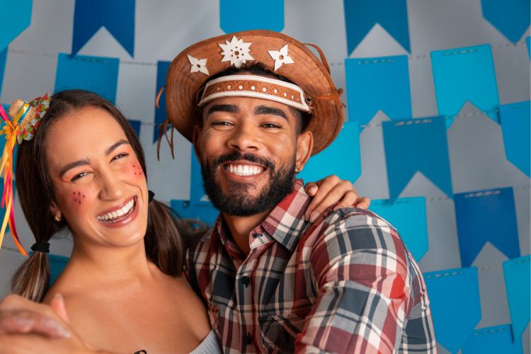 Mulher de pele clara usando trajes de festa junina abraçada com homem de pele morena também com trajes juninos, ambos à frente de bandeirinhas azuis penduradas