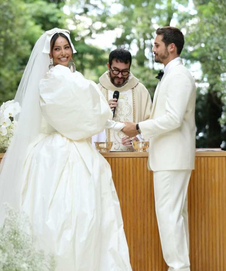 Casal Sabrina Sato e Nicolas Prattes sorridente troca votos durante cerimônia ao ar livre. Noiva em vestido volumoso e véu longo, enquanto o noivo usa terno branco elegante. Ambiente natural com padre ao centro simboliza a sofisticação dos casamentos de famosos 2025. 
