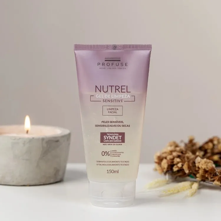 A imagem exibe um sabonete para pele sensível, o "PROFUSE NUTREL GEL DE LIMPEZA SENSITIVE", em um frasco lilás. Destaca "Limpeza Facial para peles sensíveis, sensibilizadas ou secas" e "Tecnologia Syndet". A vela acesa e galhos secos no fundo criam uma atmosfera acolhedora e natural para o produto de 150ml.







