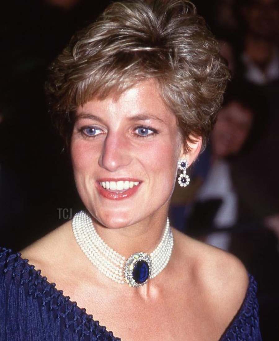 Foto de lady Di em um local com pessoas. Ela está de frente e usa gargantilha de pérolas e safira e brincos de pérola e diamantes. Atrás dela, é possível ver a silhueta de outras pessoas.