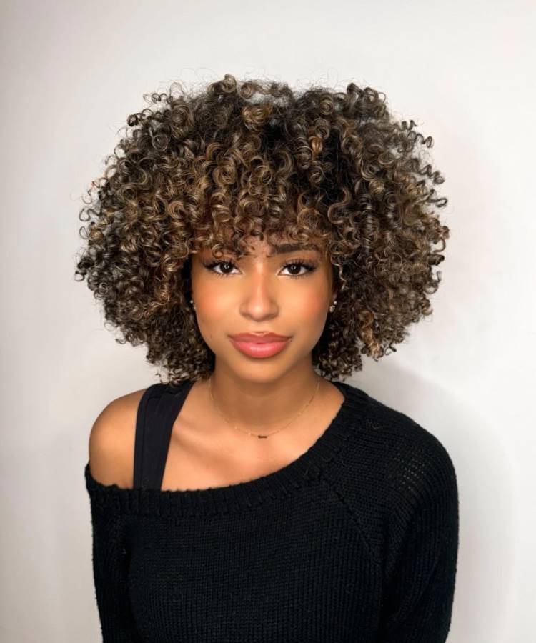 A imagem apresenta uma mulher jovem com cabelos cacheados curtos e volumosos, com mechas claras. Ela usa uma blusa preta de um ombro só e um colar discreto. A mulher olha para a frente com uma expressão suave e os lábios rosados.








