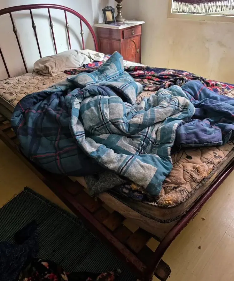 Quarto simples com cama desarrumada, colchão gasto e roupas de cama em desalinho. Cobertores coloridos e lençol amassado cobrem parcialmente a estrutura metálica do estrado. Ao lado, mesa de cabeceira de madeira com luminária. Parede mostra sinais de desgaste.