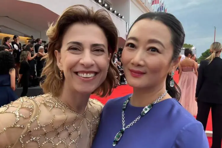 Fernanda Montenegro e Zhao posam sorridentes em tapete vermelho de festival de cinema. Brasileira veste vestido de Giorgio Armani, bege bordado com transparência, a outra usa traje azul elegante com colar de pedras verdes. Ao fundo, fotógrafos, convidados e bandeiras completam o ambiente glamouroso.