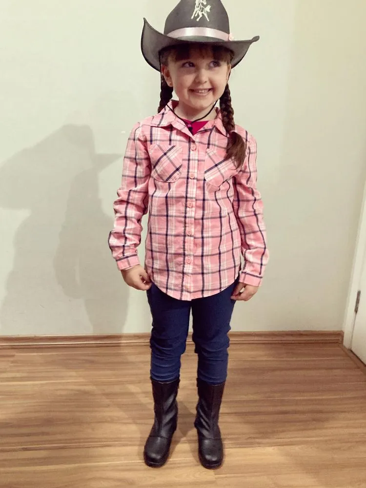 Menina de pele clara com chapéu country, camisa xadrez rosa e calça 