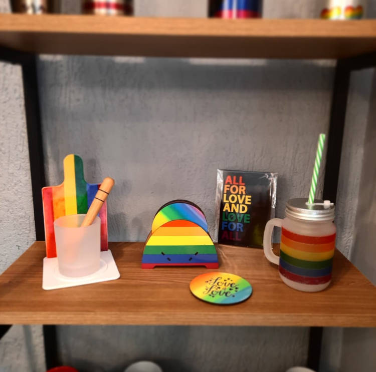 Prateleira de madeira com itens decorativos nas cores da bandeira LGBT, como caneca, porta copo, porta guarnapo e tábua