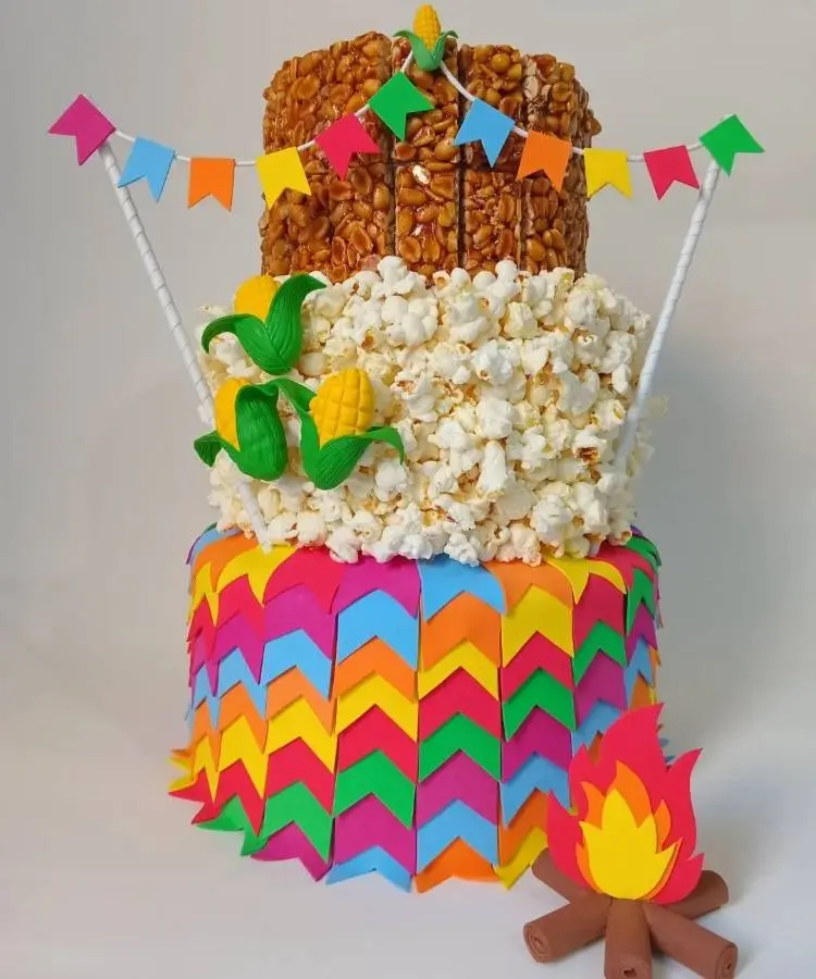 Um bolo cenográfico de três andares para festa Junina. Os andares simulam doce de amendoim, pipoca e decoração colorida, com bandeirinhas e enfeites de milho e fogueira.