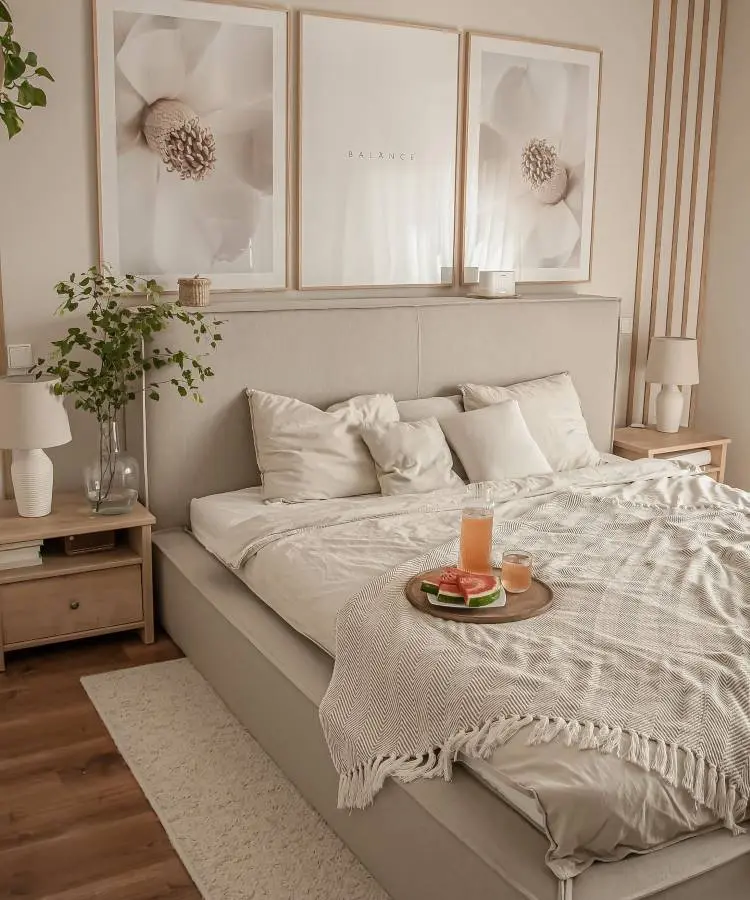 Dormitório para marido e mulher, com 3 quadros neutros acima da cabeceira estofada cinza, com mesas de canto e abajures + tapete felpudo e decor com plantas