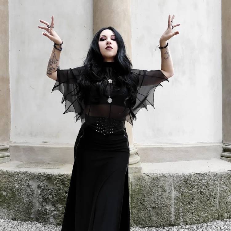 A imagem retrata uma mulher com cabelos escuros e maquiagem marcante, vestindo uma roupa preta com mangas que lembram asas de morcego. Ela está em uma pose dramática, com os braços erguidos. O cenário é de pedra e colunas, adicionando um toque gótico. Capas de Halloween são destacadas no estilo sombrio e elegante da fantasia.


