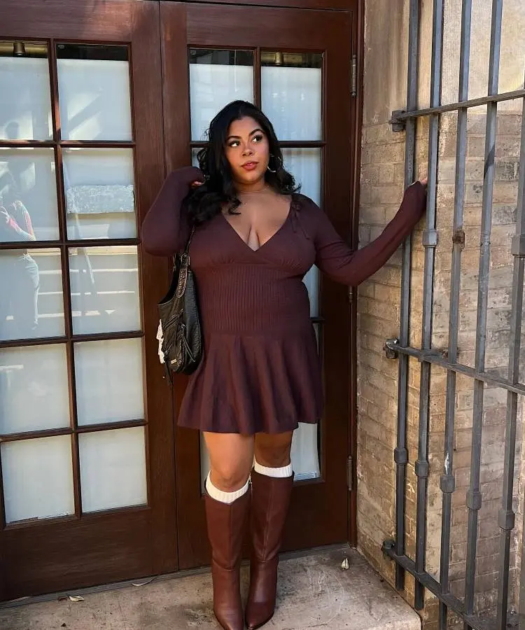 Uma mulher com cabelo escuro e ondulado veste um vestido de tricô marrom escuro com decote em V e saia rodada. Ela usa botas marrons de cano alto com meias brancas aparecendo na parte superior. A mulher está em frente a uma porta de madeira escura e uma grade.