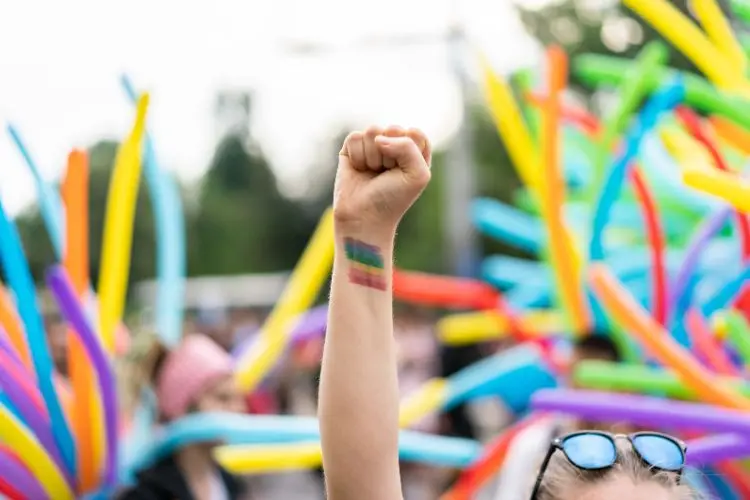 Pessoa de pele branca erguendo o punho fechado com uma pintura da bandeira LGBT em uma manifestação