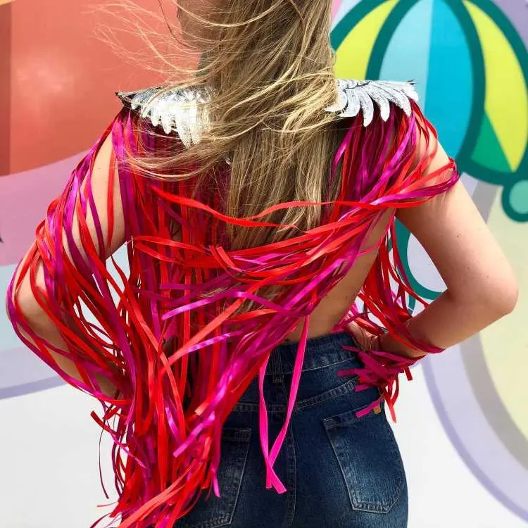 Uma mulher de costas veste ombreiras prateadas com franjas longas e coloridas em tons de rosa e vermelho. Seu cabelo loiro está solto, e ela usa calça jeans escura. Ao fundo, há um painel colorido com guarda-chuvas.