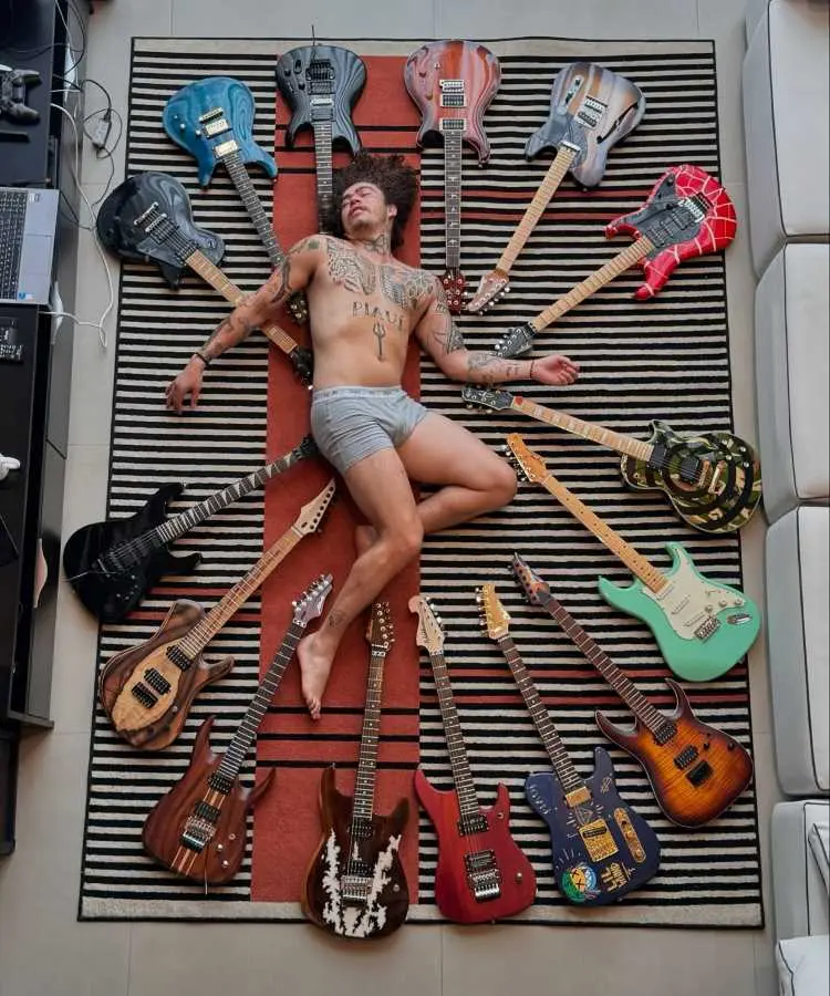 A imagem mostra Whindersson Nunes deitado em um tapete listrado, cercado por várias guitarras de diferentes cores e estilos. Ele está usando apenas uma cueca cinza, com tatuagens visíveis no corpo e expressão descontraída.