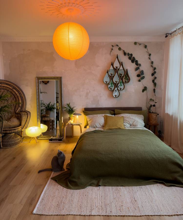 Quarto com decoração acolhedora em tons quentes, cama com colcha verde-oliva, luz suave de luminária pendente laranja, planta trepadeira decorando a parede, espelho grande ao lado, cadeira de vime, cortinas leves e gato sentado no tapete.





