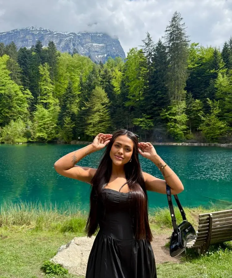 Camila Loures de cabelos escuros e longos, vestindo um vestido preto, posa em frente a um deslumbrante lago azul-turquesa. Atrás dela, uma densa floresta de pinheiros sobe em direção a majestosas montanhas sob um céu nublado. Ela sorri para a câmera, com as mãos perto da cabeça.