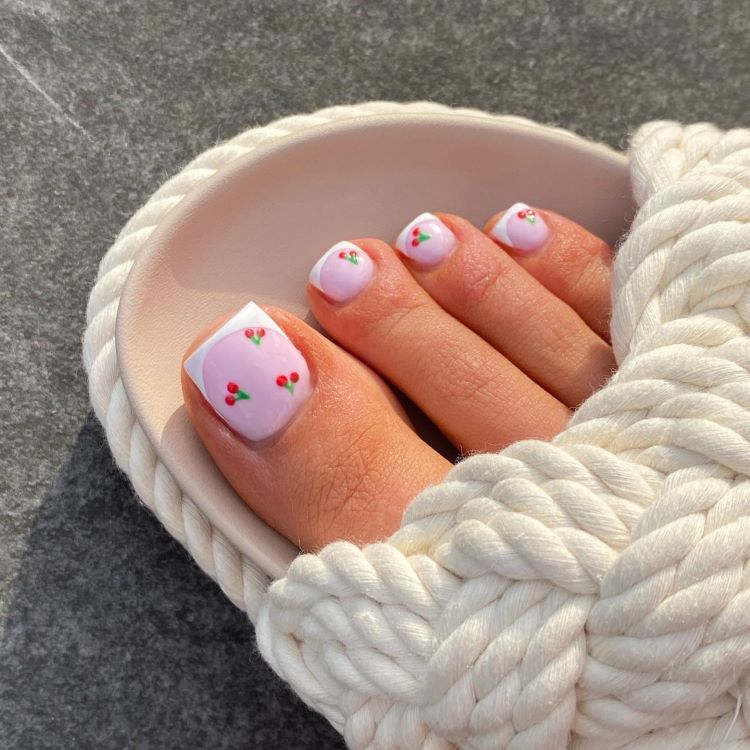 Pés em destaque com unhas decoradas em esmalte rosa claro, desenhos de pequenas cerejas e acabamento em francesinha branca. Sandália de tecido trançado branco contrasta com o fundo de pedra cinza iluminado pelo sol, criando um estilo delicado e charmoso.





