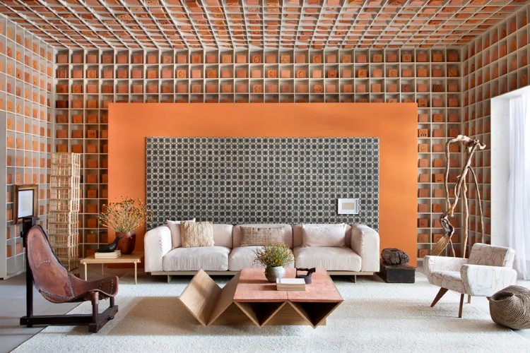 A imagem mostra uma sala de estar com estilo moderno, paredes de tijolos aparentes laranjas, sofá bege, poltronas marrons e brancas, mesa de centro geométrica em madeira e decoração minimalista.

