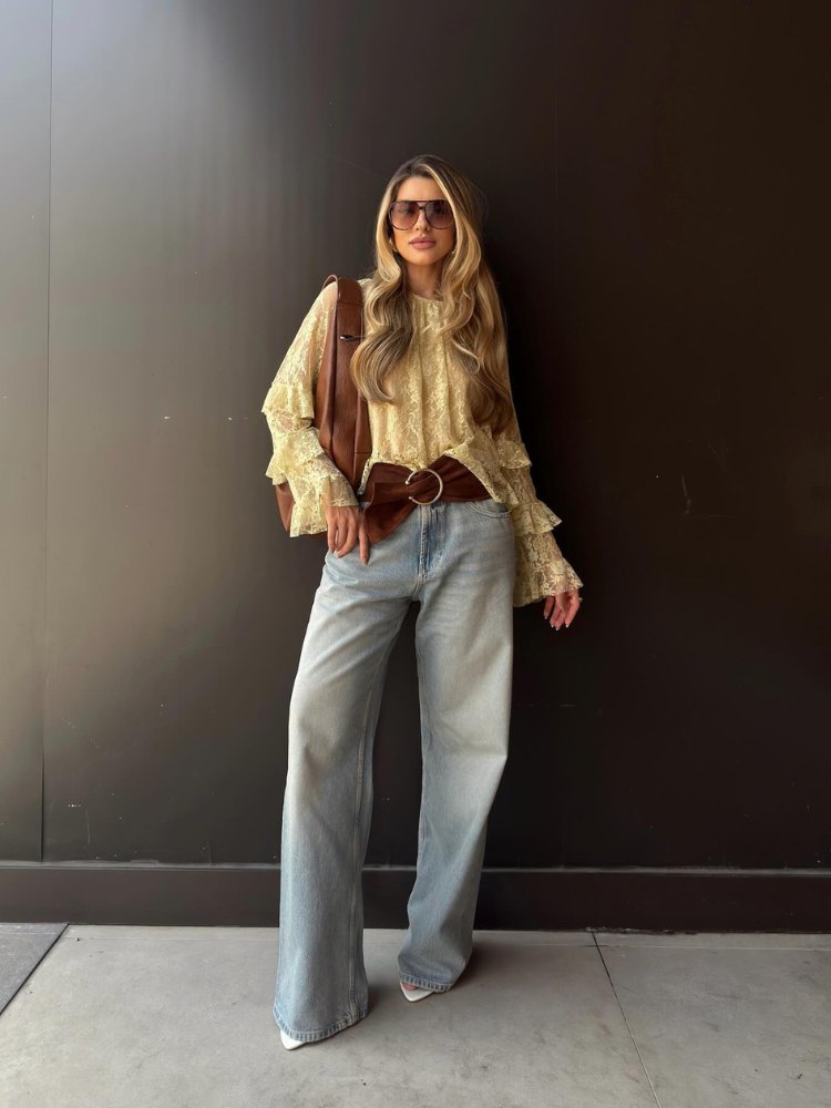 Mulher de pele clara com camisa amarela de babados, cinto marrom de camurça, calça jeans e bolsa marrom 