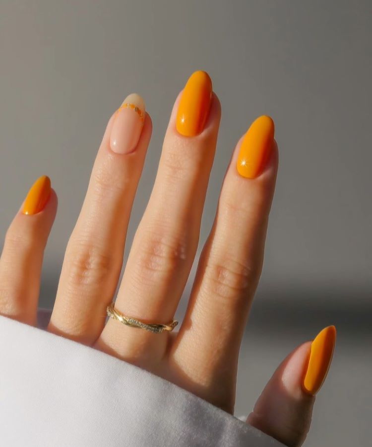 A imagem mostra uma mão com unhas pintadas em um tom vibrante de laranja, exceto uma que está em um tom natural com uma linha dourada na ponta, criando um destaque delicado. Este design é um exemplo de unha decorada simples, que combina um toque de cor com um detalhe minimalista. A mão está adornada com um anel dourado e veste uma camisa branca, destacando ainda mais a simplicidade e elegância do estilo.