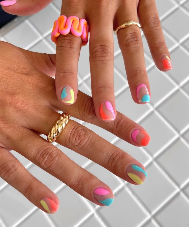 Mãos com estilos de unhas curtas, decoradas com esmalte nude como base e desenhos ondulados em tons vibrantes de rosa, laranja, amarelo, azul e verde. Dedos adornados com anéis dourados e coloridos. Fundo com padrão geométrico branco destacado.