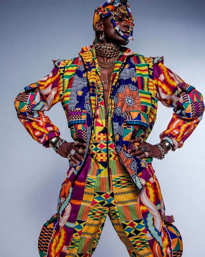 Modelo masculino com terno colorido e estampado, e acessórios no pescoço e cabeça.