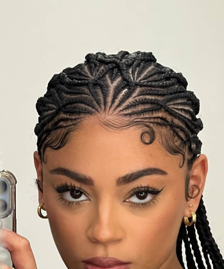 Mulher com tranças finas estilizadas em padrão geométrico rente ao couro cabeludo. Baby hairs modelados em curvas decorativas na testa. Ela usa brincos de argola dourados, maquiagem com delineado marcante, pele impecável e olha diretamente para a câmera.