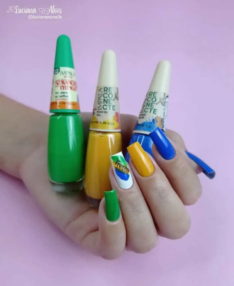 Imagem de mão com unhas decoradas para a Copa do Mundo 2022. Cores usadas, verde, amarelo, azul e branco. Polegar, azul. Indicador, azul. Médio, amarelo. Anelar, branco com cores verde, amarelo e azul com desenho preto de galho. Mínimo, verde. 