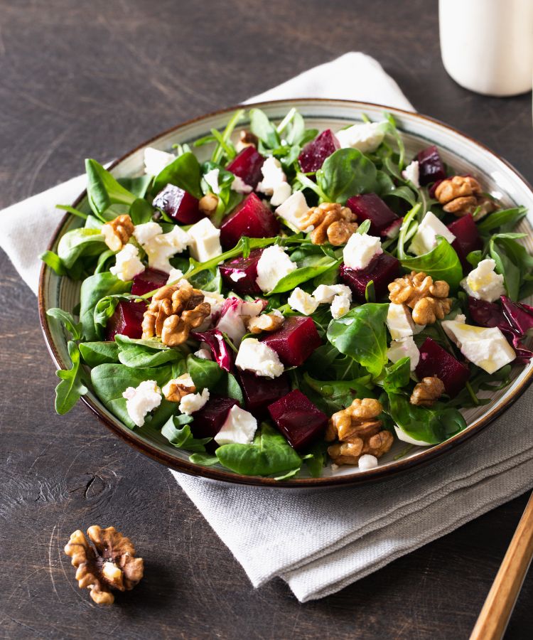 Uma salada vibrante em um prato com folhas verdes, cubos de beterraba, nozes, e queijo feta. A salada repousa sobre um guardanapo de linho em uma mesa de madeira escura.