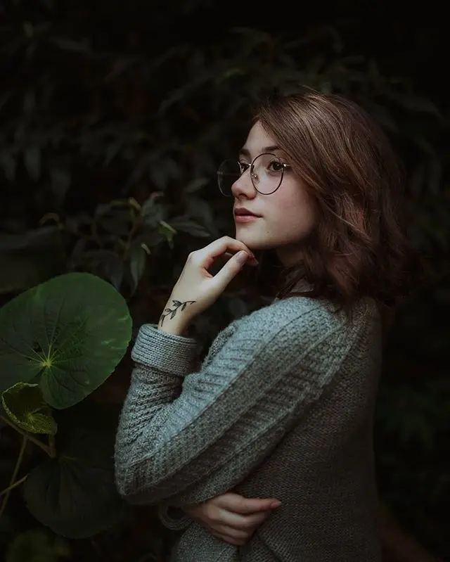 Frase para foto pensativa mulher branca com tatuagem na mão, usa óculos de grau e veste blusa de tricot