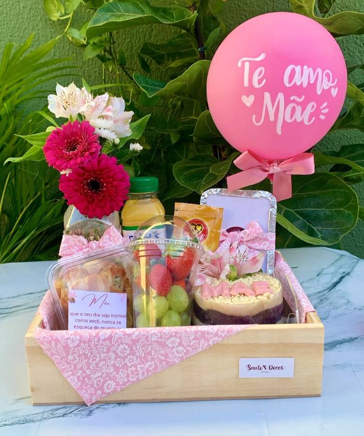 Cesta de presente para o Dia das Mães com balão rosa "Te amo Mãe", flores, suco, frutas e bolo decorado. Inclui mensagem afetiva e lanches, dispostos em bandeja de madeira com tecido estampado rosa. Fundo com plantas verdes.
