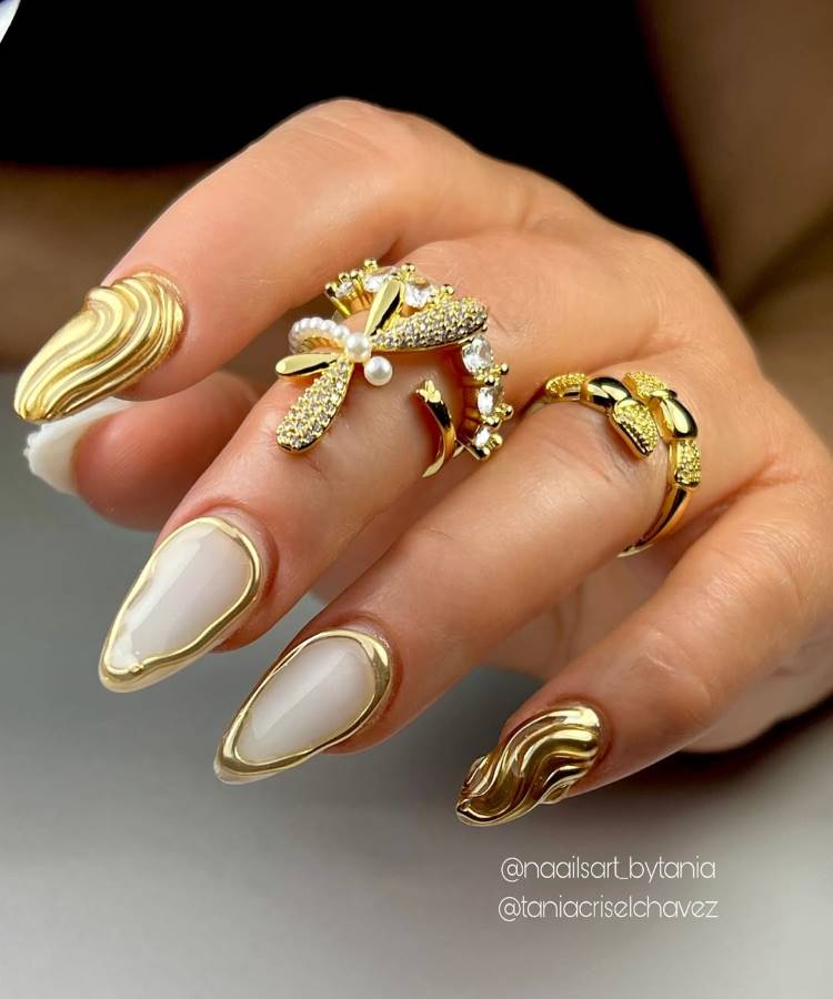 A imagem destaca unhas decoradas elegantemente em tons dourados e branco perolado, com detalhes metálicos. Anéis chamativos, incluindo formas de libélula cravejadas de pedras, complementam a estética luxuosa. O fundo escuro realça o brilho sofisticado das jóias e unhas.