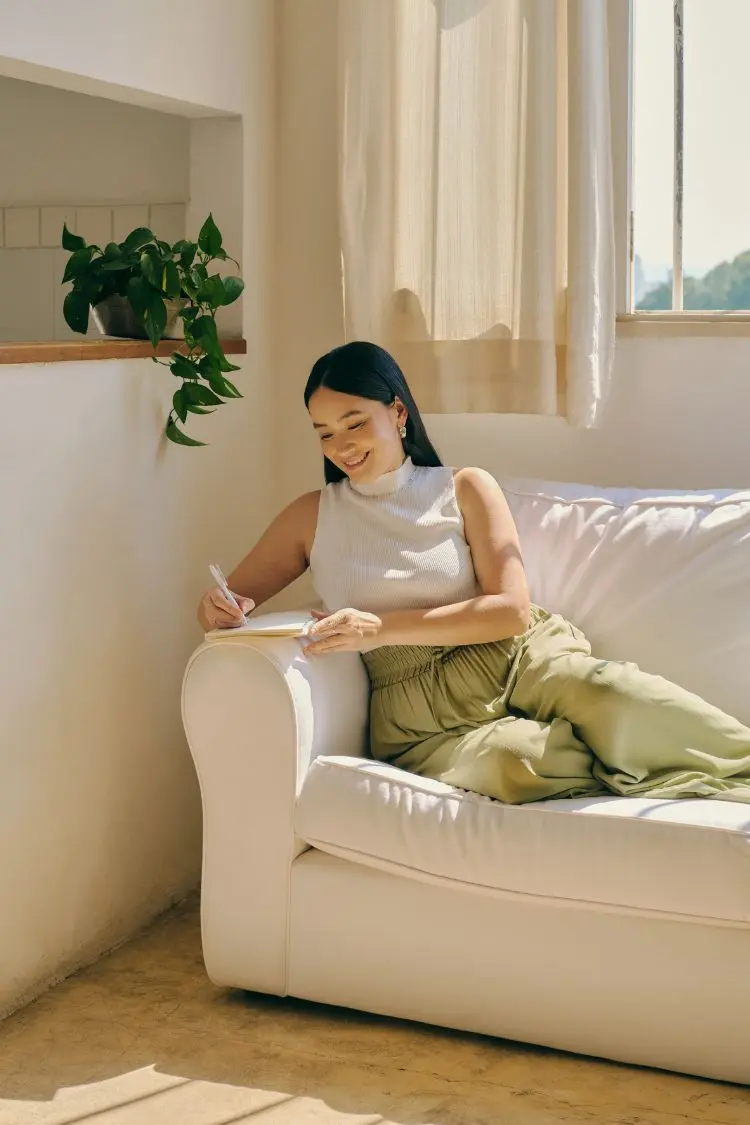 Uma mulher asiática senta-se em um sofá branco, escrevendo em um caderno com uma caneta. Ela usa blusa branca sem mangas e calças verde-oliva. A luz do sol entra pela janela. Uma planta verde está visível ao fundo, criando um ambiente relaxante.







