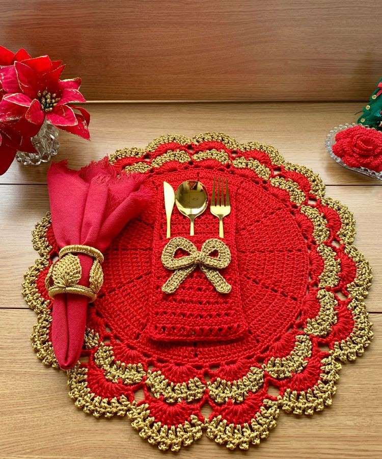 O sousplat de crochê vermelho com bordas douradas exibe um porta-talheres decorado com laço. Talheres dourados complementam a cena, ao lado de uma flor vermelha decorativa.


