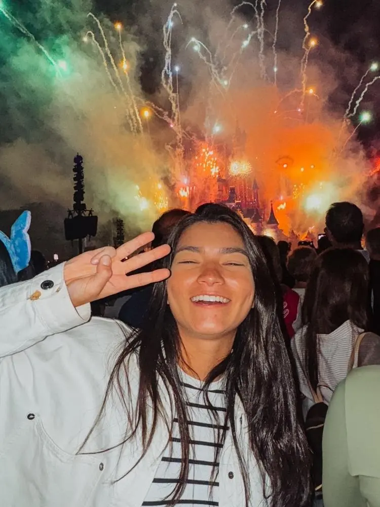 Lara Santana usando blusa branca de listra, jaqueta branca, fazendo o número 3 com os dedos e fogos da Disney ao fundo 