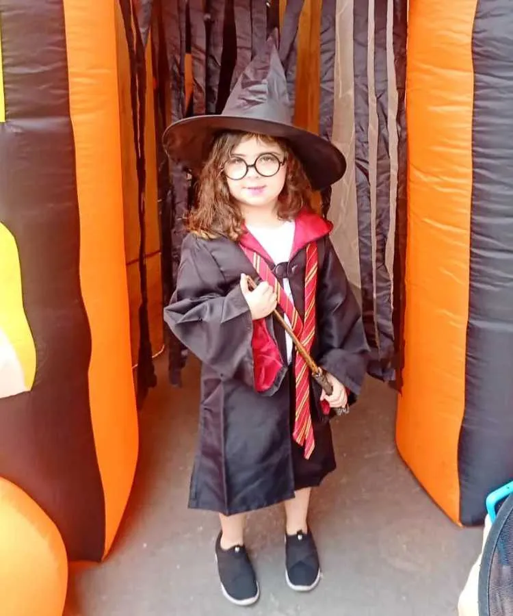 Garota fantasiada para o Halloween Harry Potter: Cores predominantes: vermelho, preto. Usa: capa com gravata, tênis, chapéu pontudo, segura varinha mágica
