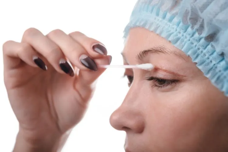 Mulher cuidando da cirurgia de blefaroplastia, usando touca e passando cotonete na cicatriz nas pálpebras