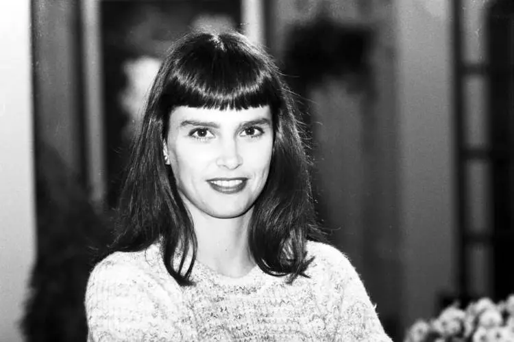 Mulher jovem de cabelos castanhos lisos com franja, sorrindo suavemente. Veste um suéter claro. A imagem é em preto e branco, com fundo desfocado mostrando formas sutis e flores. Expressão transmite serenidade e confiança.